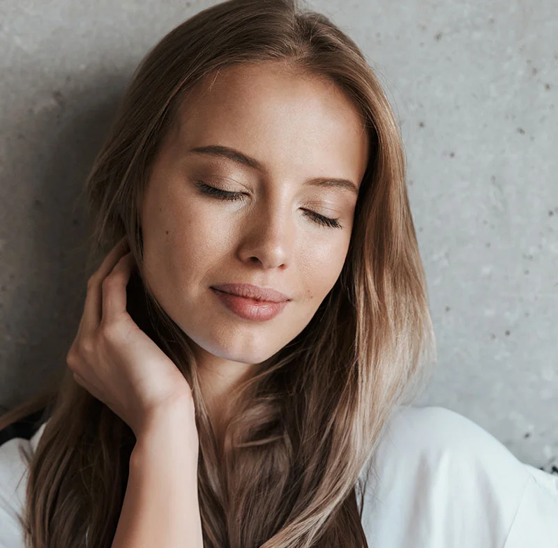 A blonde woman in a white shirt, against a grey wall, with her eyes closed - Micro Laser Peel in Red Bank, NJ