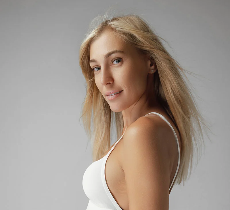 A blonde woman in a white tank top, smiling with her hair slightly flowing in the wind, against a grey background - Upneeq in Red Bank, NJ